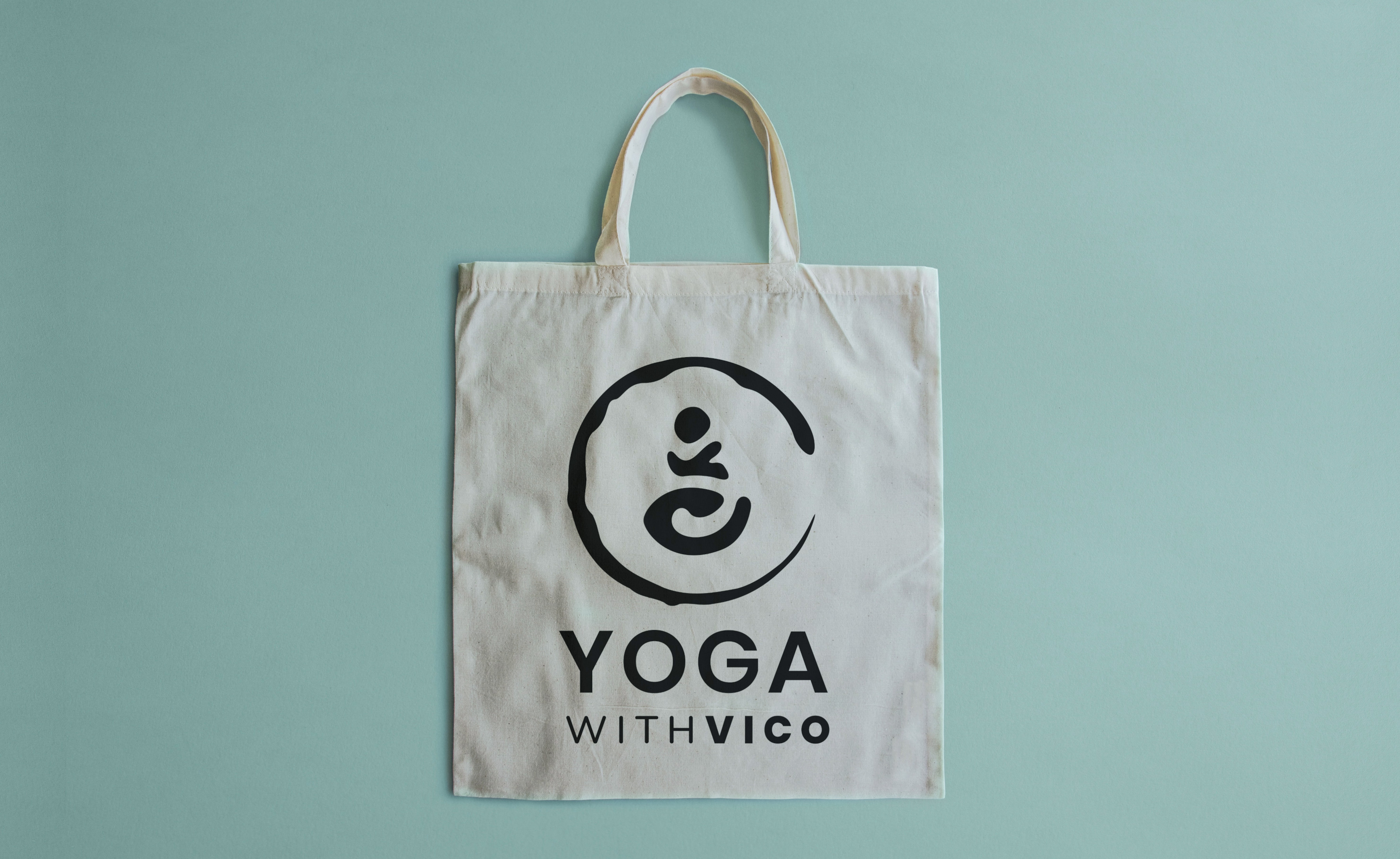 Yoga with Vico's square logo design shown on a natural fibre tote bag. Background is plain and light aquamarine colored.
